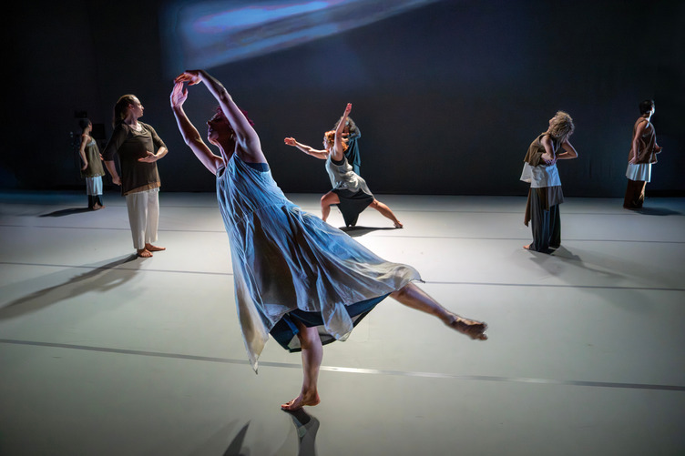 A woman dancer wearing a flowing dress in the foreground, lifts her arms in a circle above her head while extending her leg in a low arabesque.